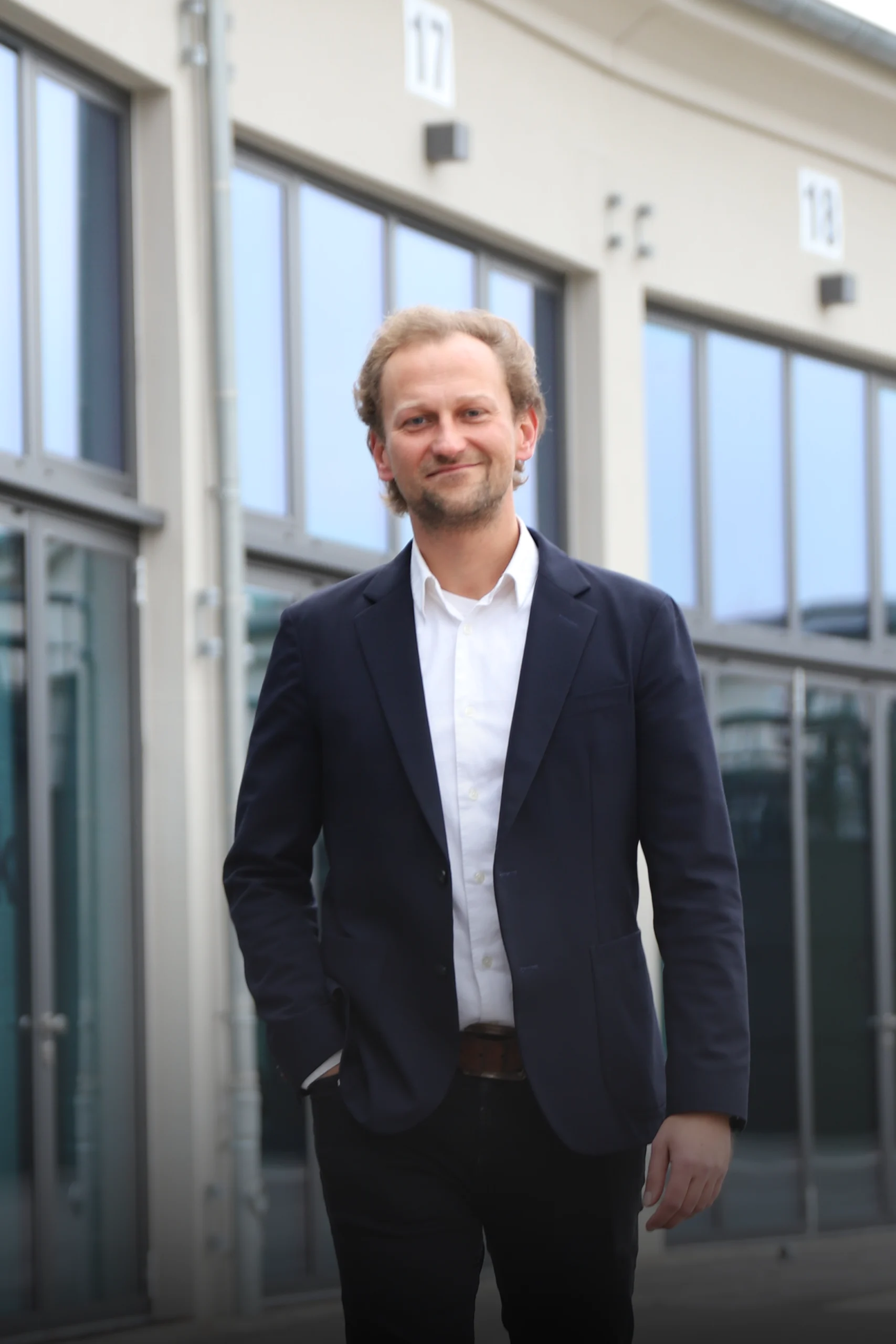 Eine Person im Anzug lächelt vor modernen Glasgebäuden in einer urbanen Umgebung.