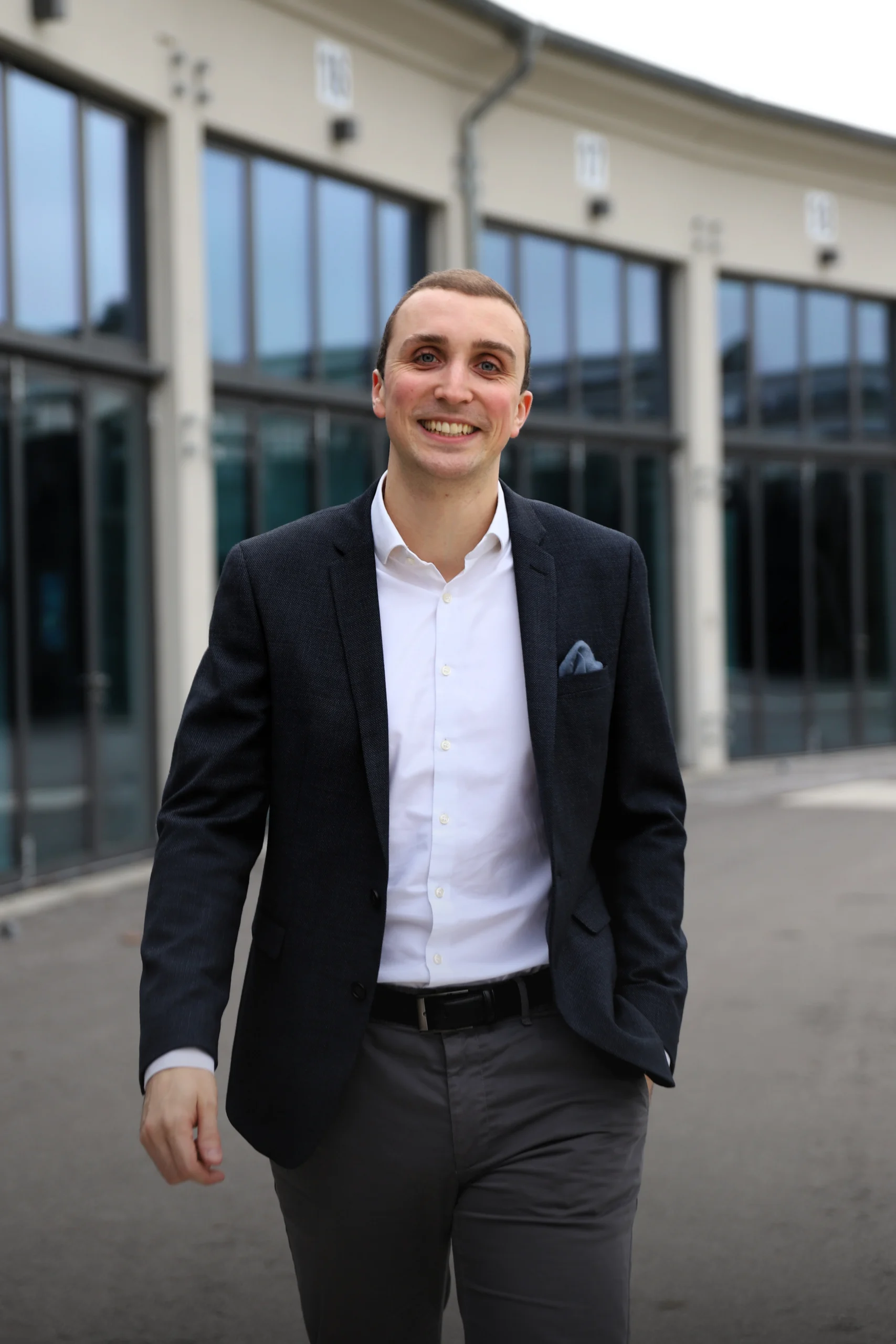 Ein lächelnder Mann im Anzug geht vor modernen Gebäuden mit Glasfenstern auf den Betrachter zu.