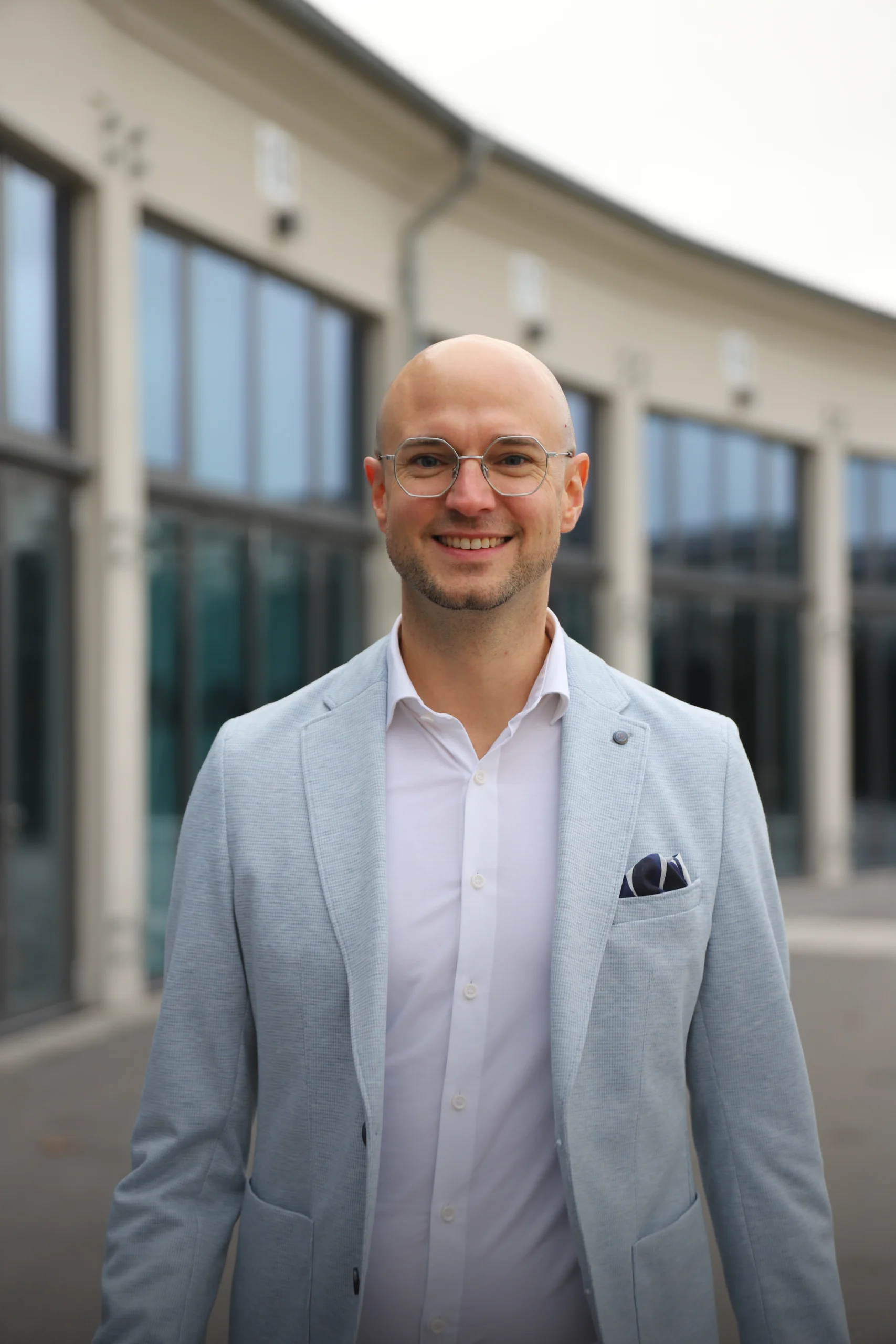 Eine lächelnde Person in hellblauem Sakko steht vor modernen Gebäuden mit Glasfenstern.