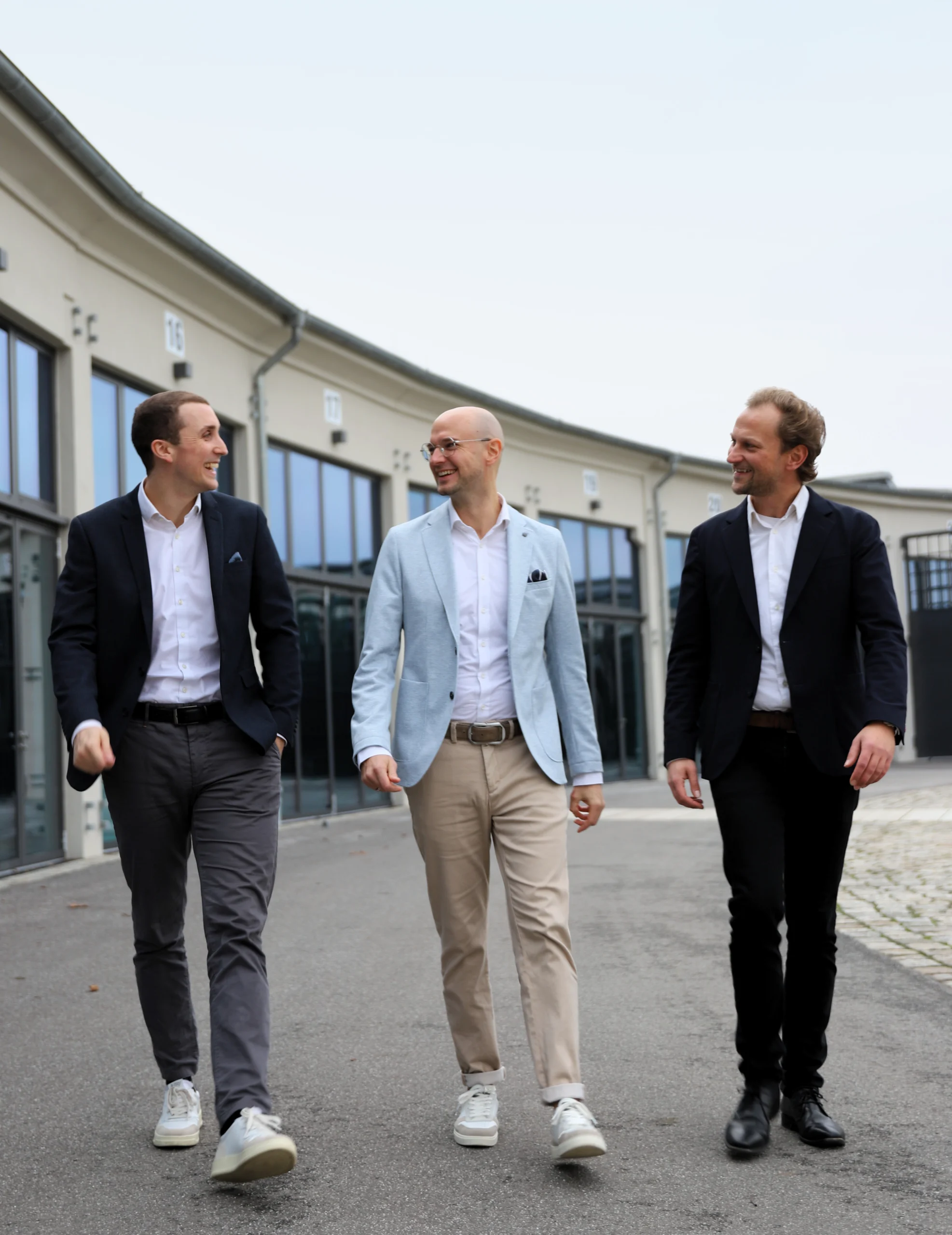 Drei Männer in Business-Kleidung gehen lächelnd auf einer urbanen Straße, moderne Gebäude im Hintergrund.