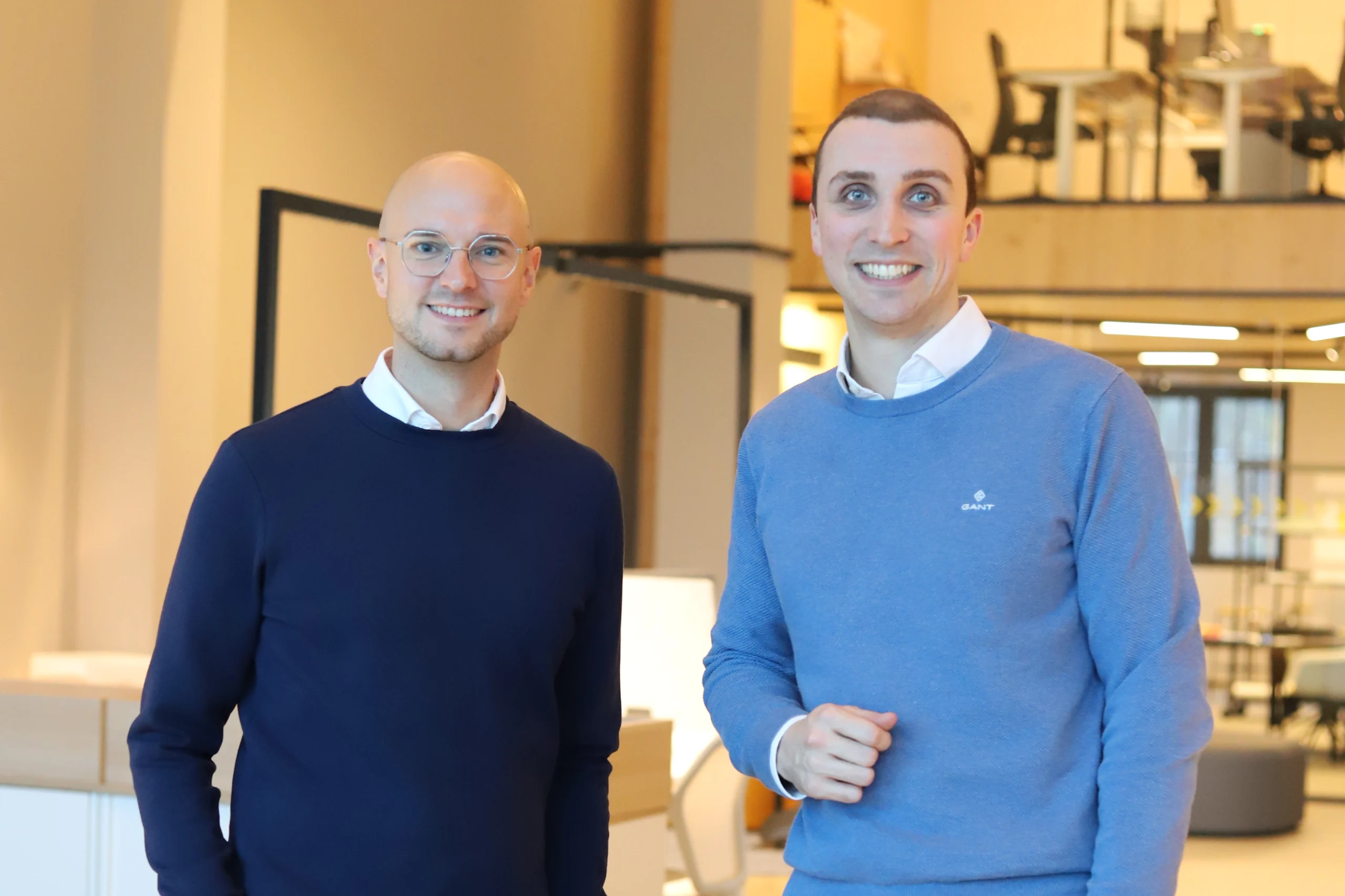 Zwei lächelnde Personen in blauen Pullovern stehen in einem modernen, gut beleuchteten Innenraum.