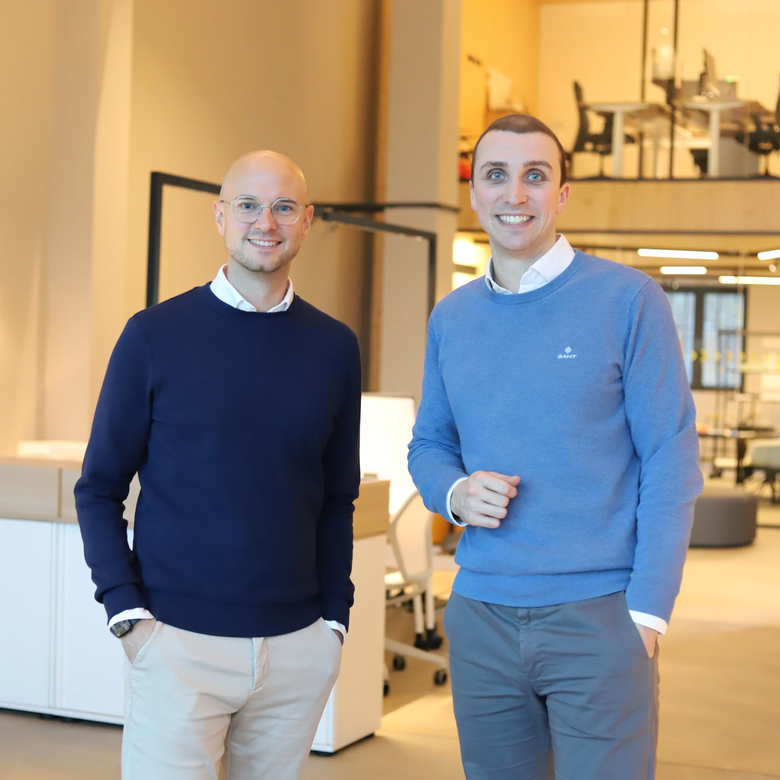 Zwei Männer in Pullovern lächeln in einem modernen, lichtdurchfluteten Büro mit minimalistischer Einrichtung.