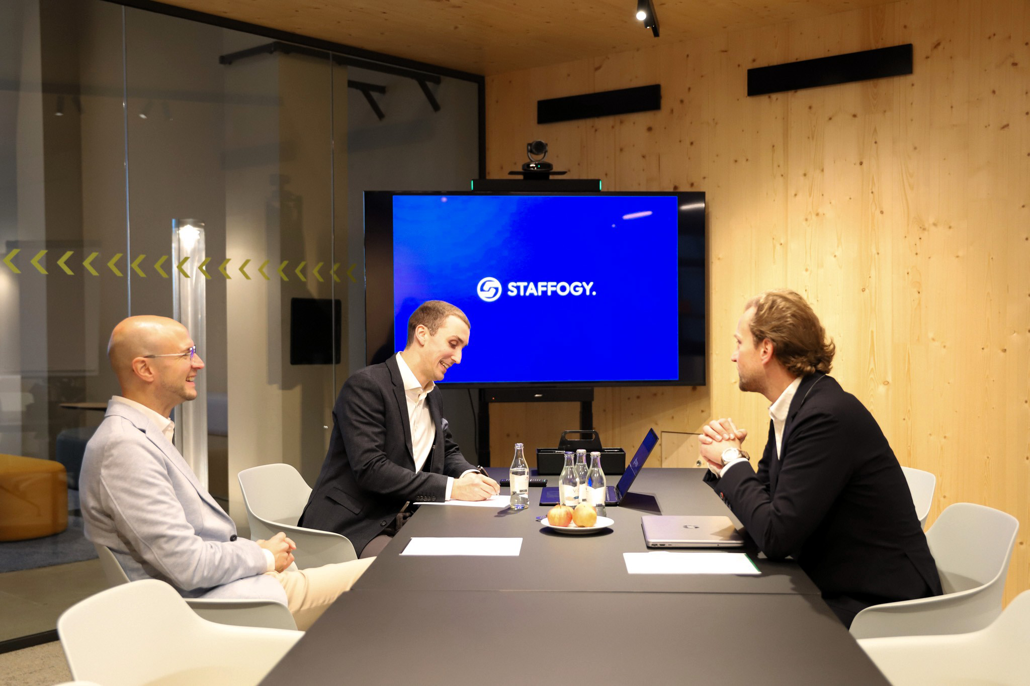 Drei Männer in Business-Kleidung in einem modernen Konferenzraum bei einem Meeting mit "STAFFOGY"-Logo.