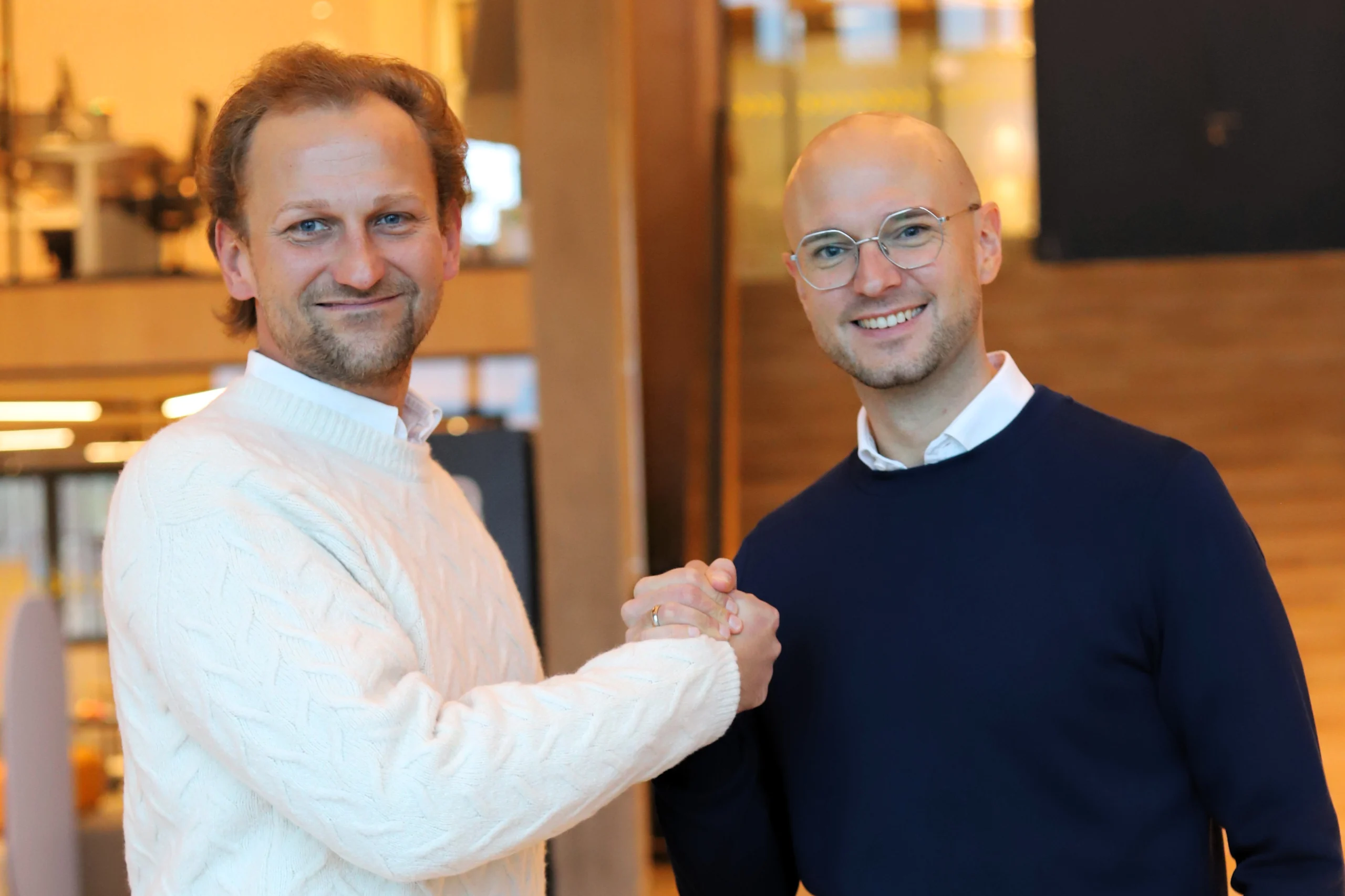 Zwei Männer lächeln und schütteln sich die Hände in einem warm beleuchteten, modernen Raum.