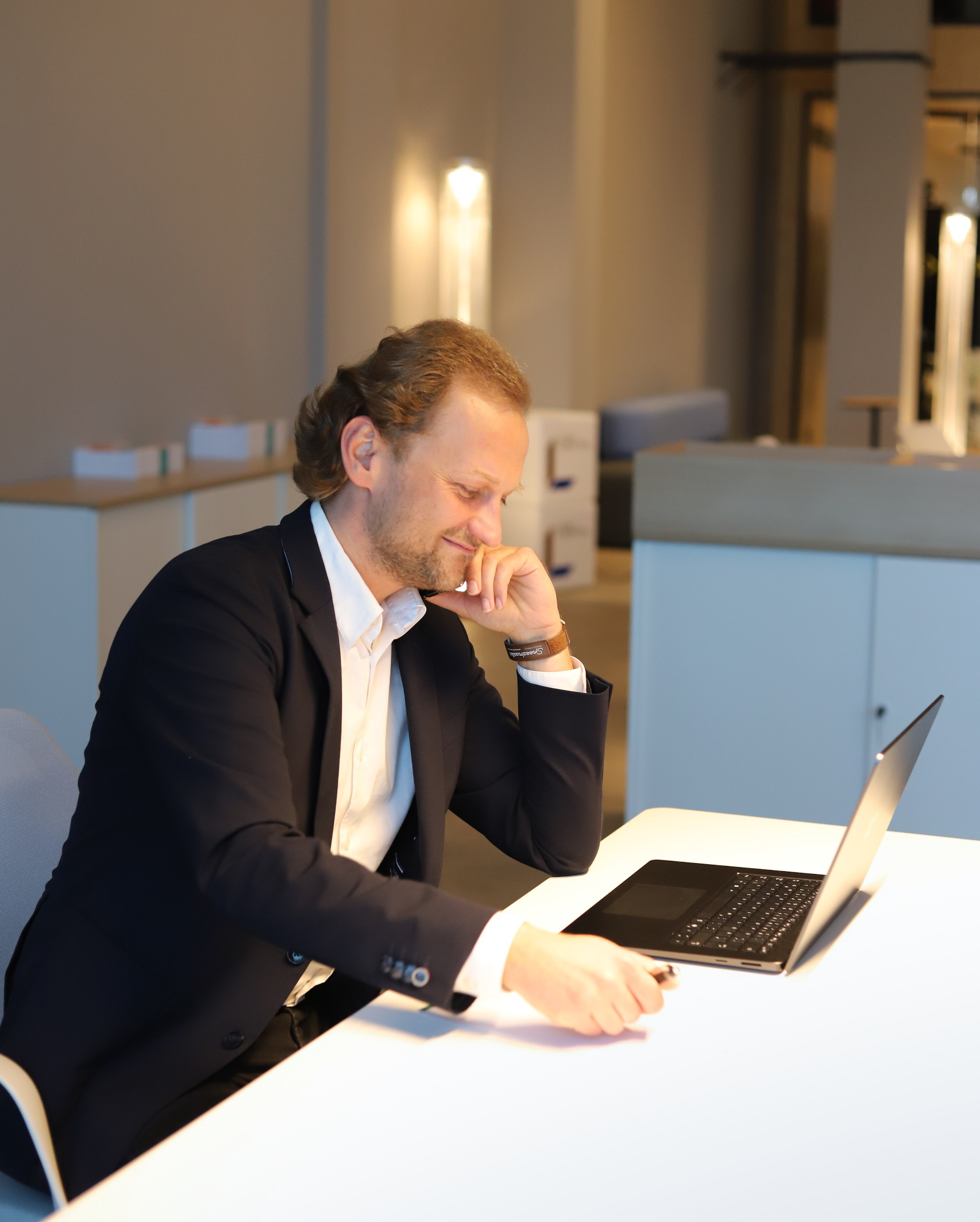 Ein Mann im Anzug sitzt nachdenklich an einem Tisch und schaut auf einen Laptop in einem modernen Büro.